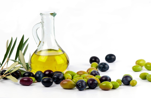 Transparent glass jug filled with golden olive oil, surrounded by a variety of fresh green and black olives, with olive branches, isolated on a white background