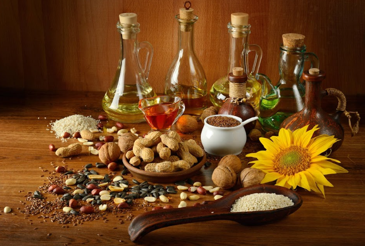 Variety of glass bottles containing different oils alongside a collection of nuts, seeds, and a bright sunflower on a rich wooden surface