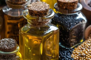 Collection of edible oils in small vintage glass jars with cork lids, surrounded by piles of various seeds and legumes like flax, black beans, and soybeans on a rustic wooden table.